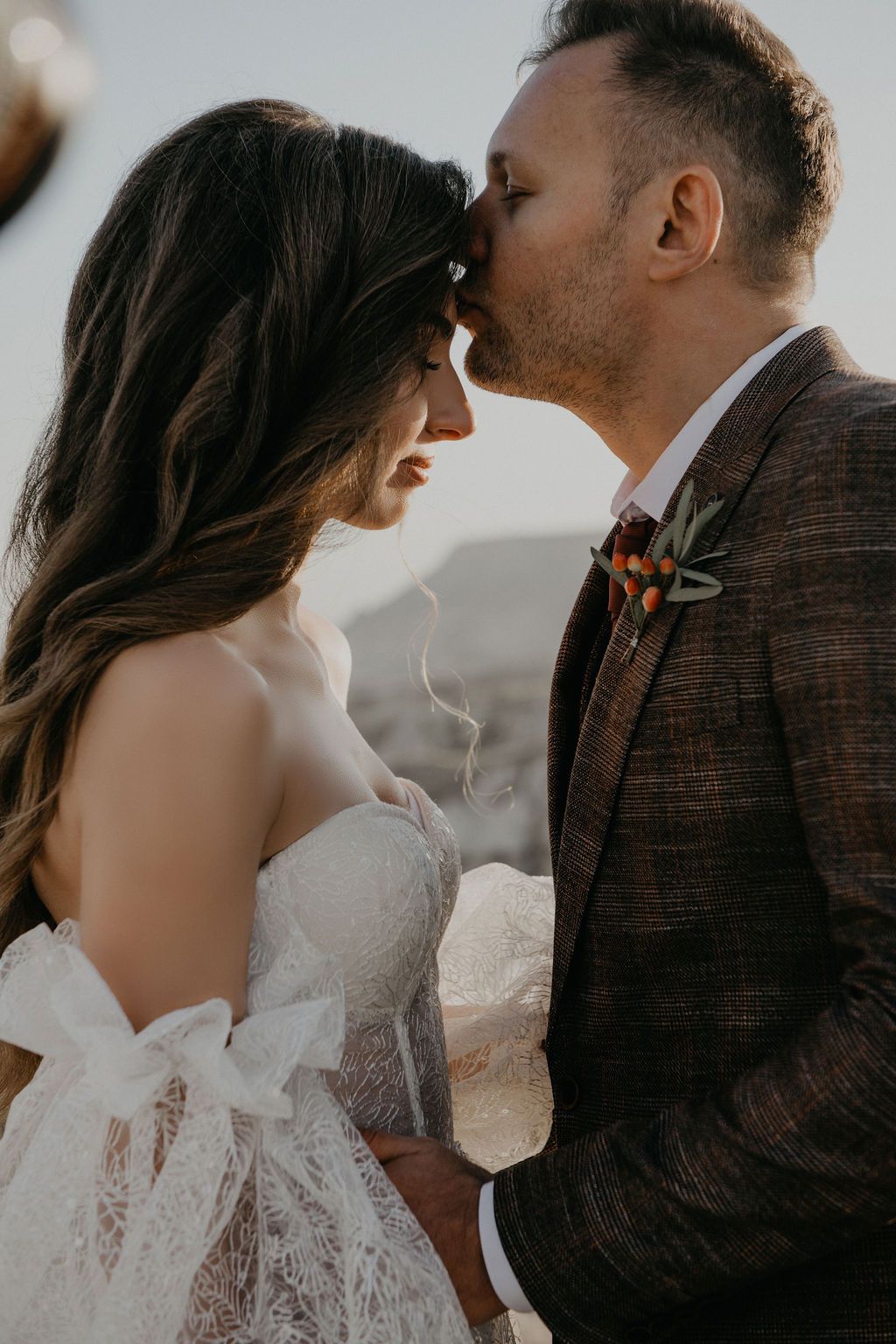 Groom in brown plaid suit tenderly kissing bride on forehead during romantic outdoor wedding photo session.