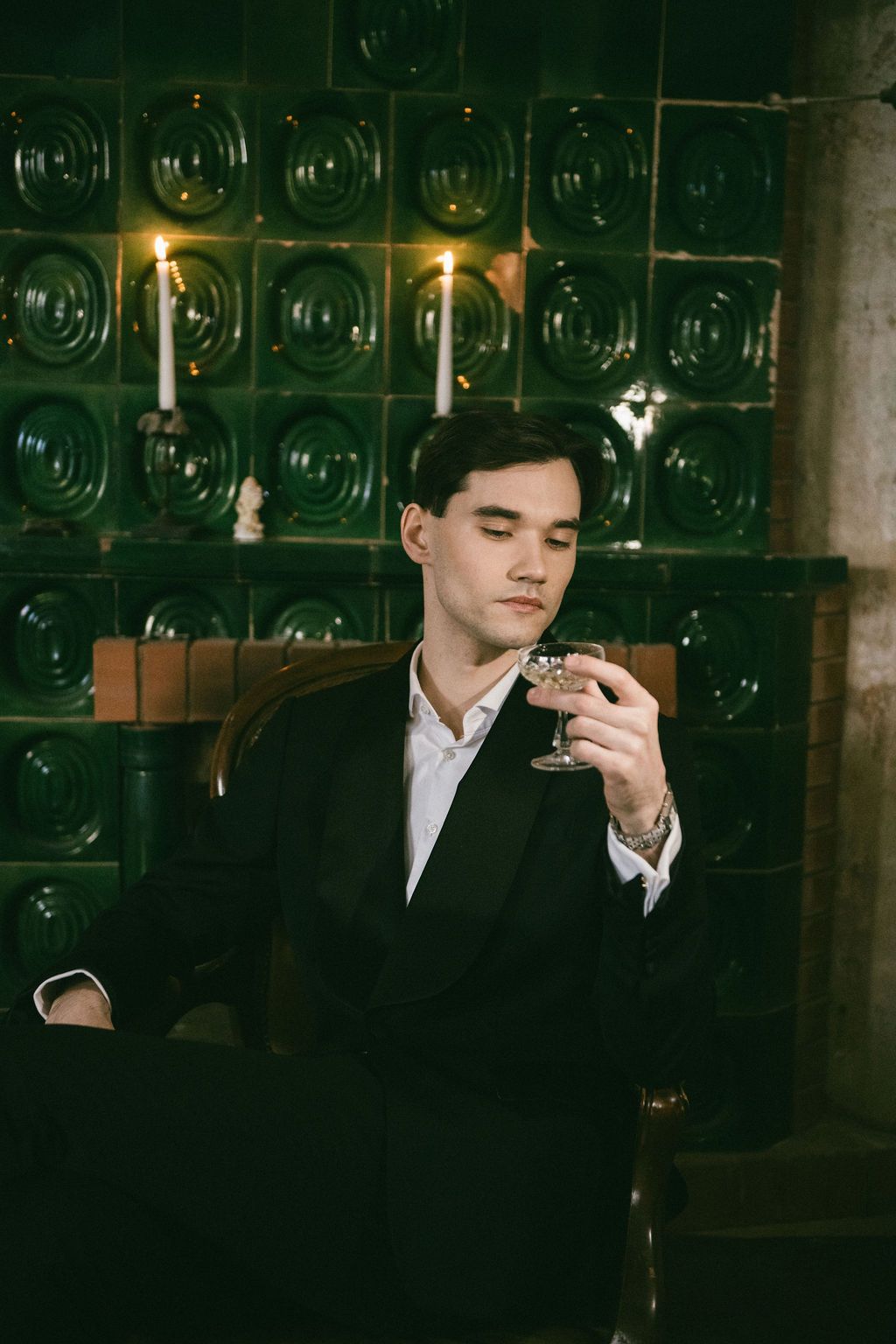 Elegant young man in black suit holding wine glass seated in vintage chair against green tiled wall with candles.