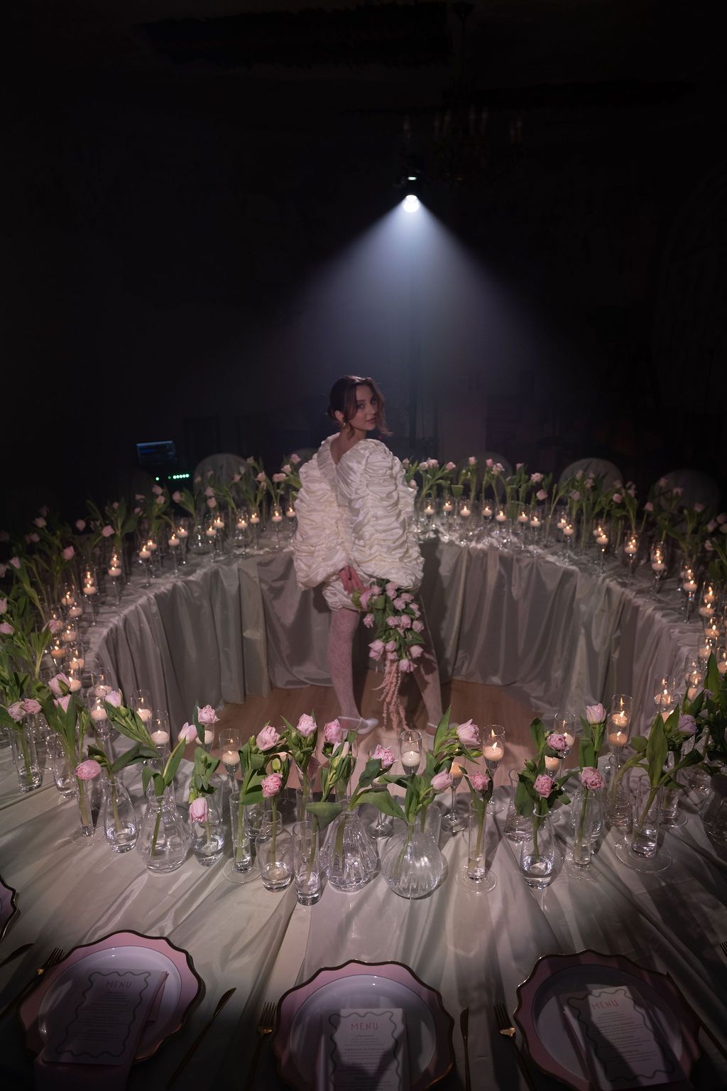 Bride in white ruffled gown surrounded by pink tulips, candles, and glass vases on a circular table in dim light.