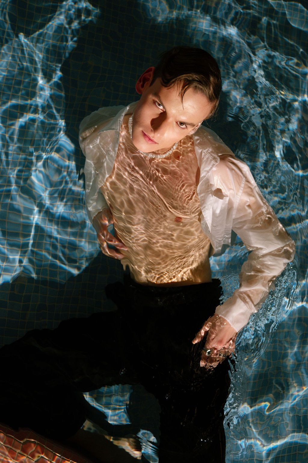 Person in wet white shirt and black pants sitting in a pool with blue rippling water reflections.