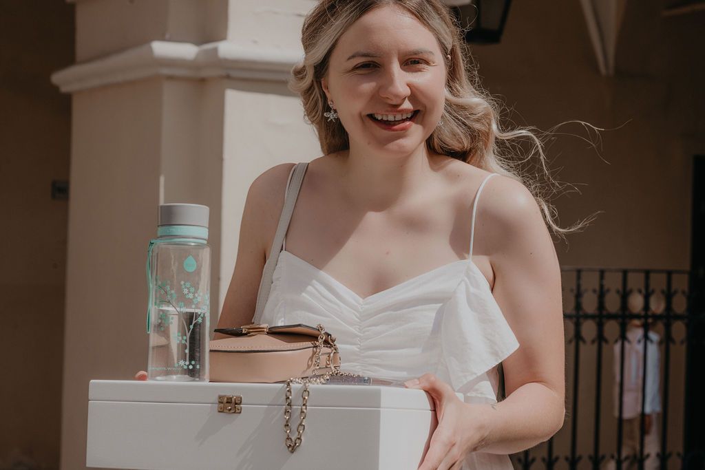 Smiling blonde bride in white dress holding a white jewelry box and water bottle outdoors.