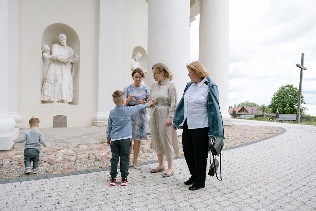 Group of women and children gathered outside a white classical church with stone statues and brick pathway.
