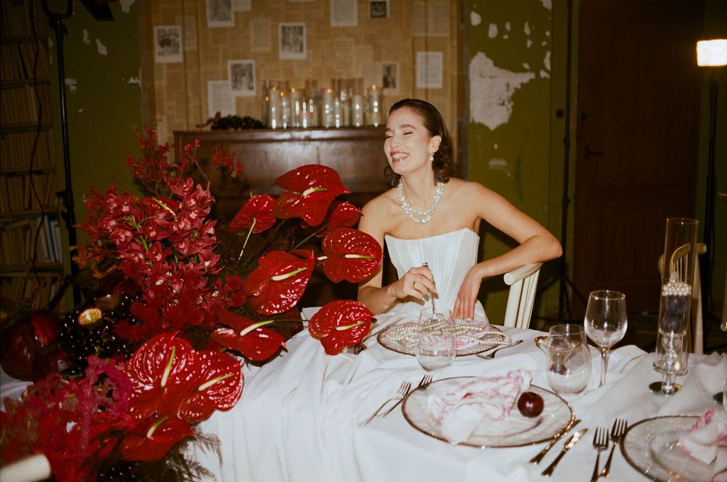 Bride in white gown seated at elegant table with red anthurium flowers in vintage-styled room.