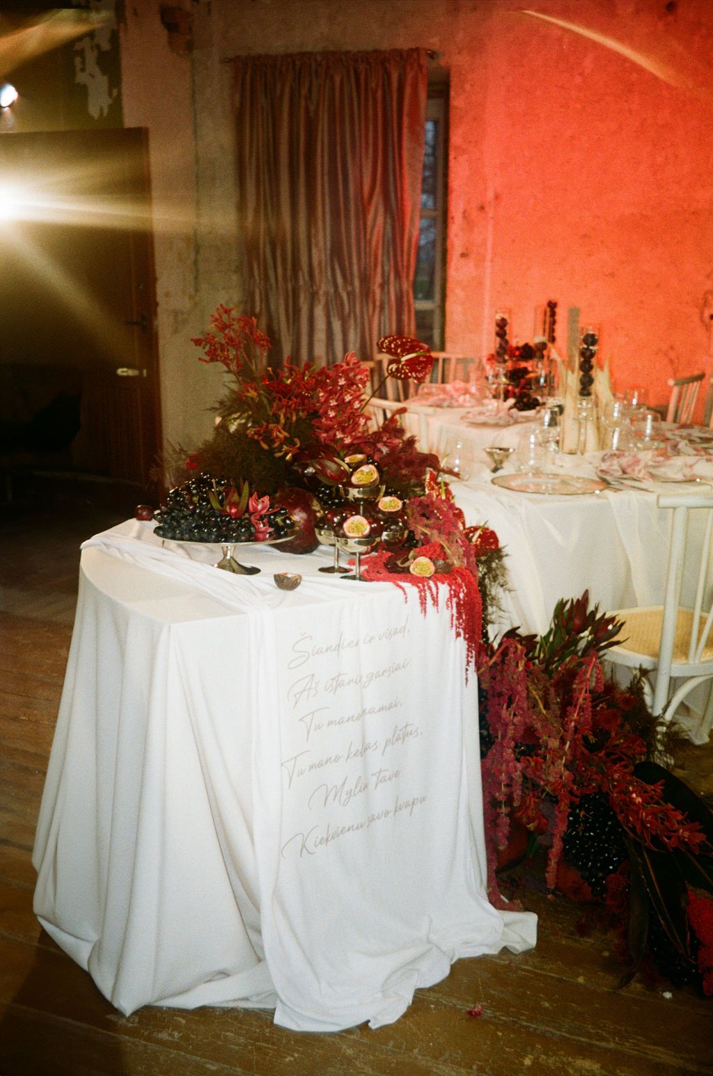 Elegant wedding reception table with white linen, red floral decor, candles, and calligraphy tablecloth in a warmly lit venue.