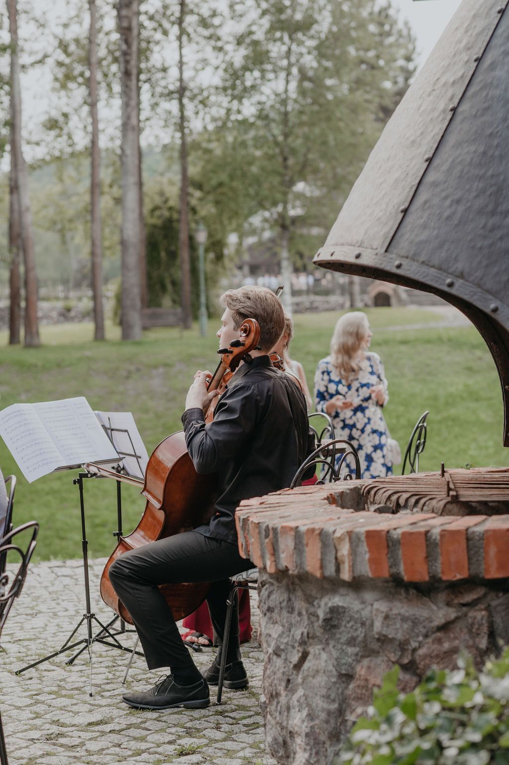 Cellist in black performing outdoors at a garden event near a stone well, with sheet music stand and trees in background.