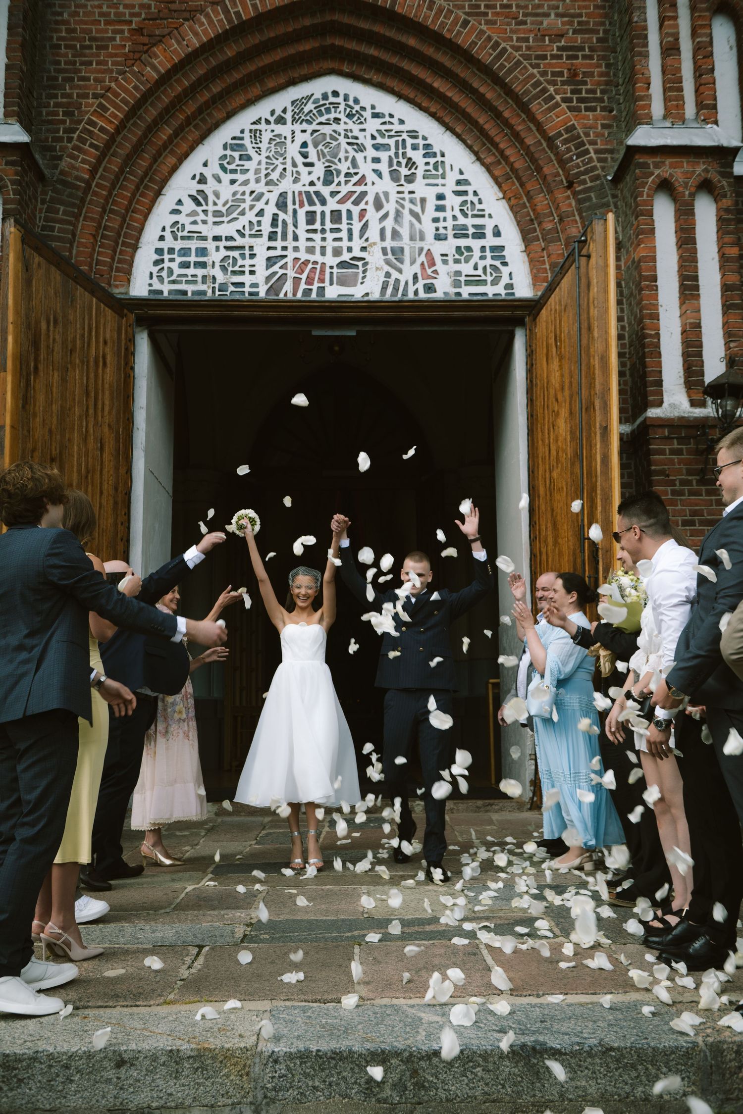 Bride in a white dress and groom in a dark suit raise their arms as guests toss white petals outside a brick church entrance.