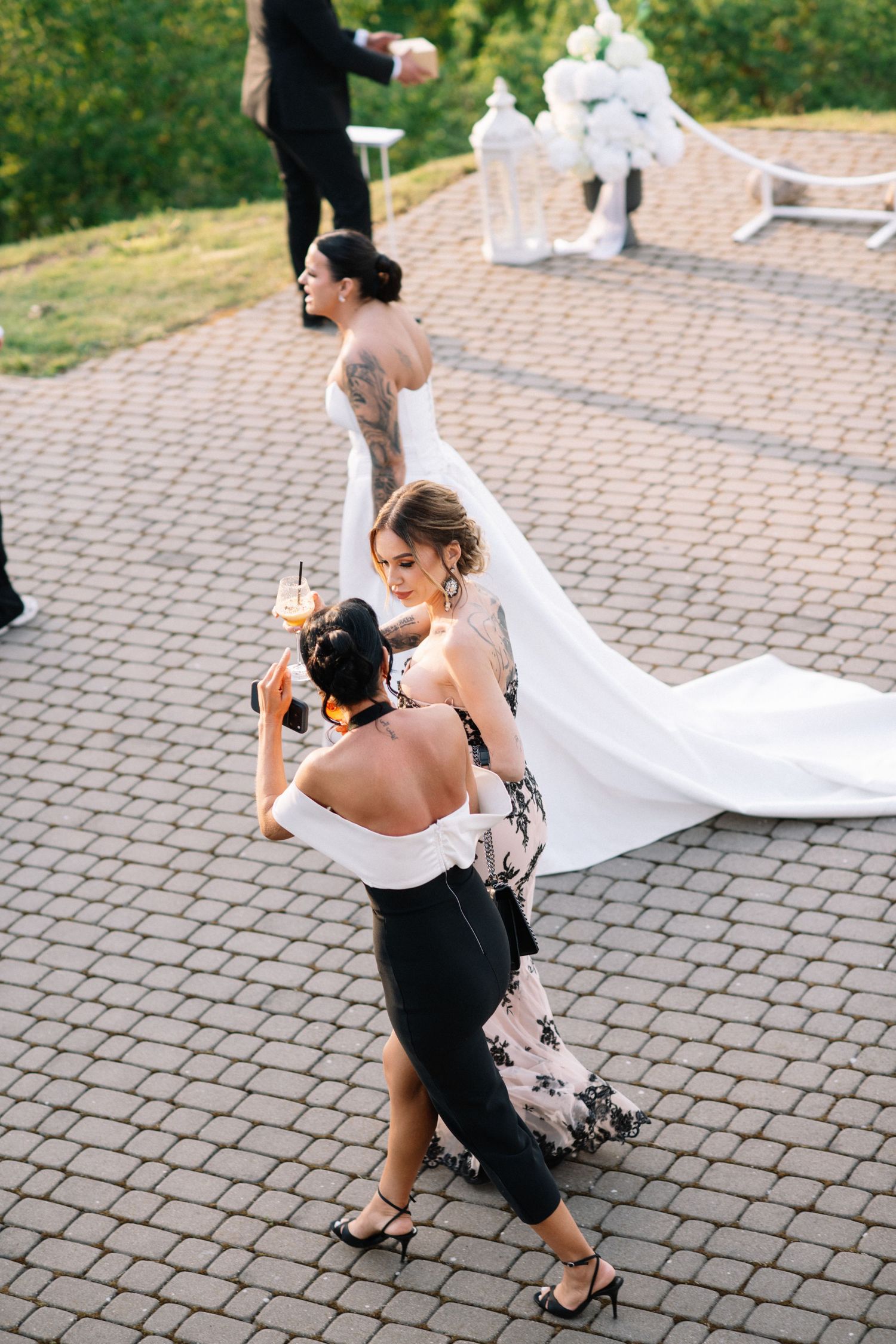 A bridesmaid in a black and white outfit adjusts her hair on a cobblestone path as a bride in a long white gown walks ahead.