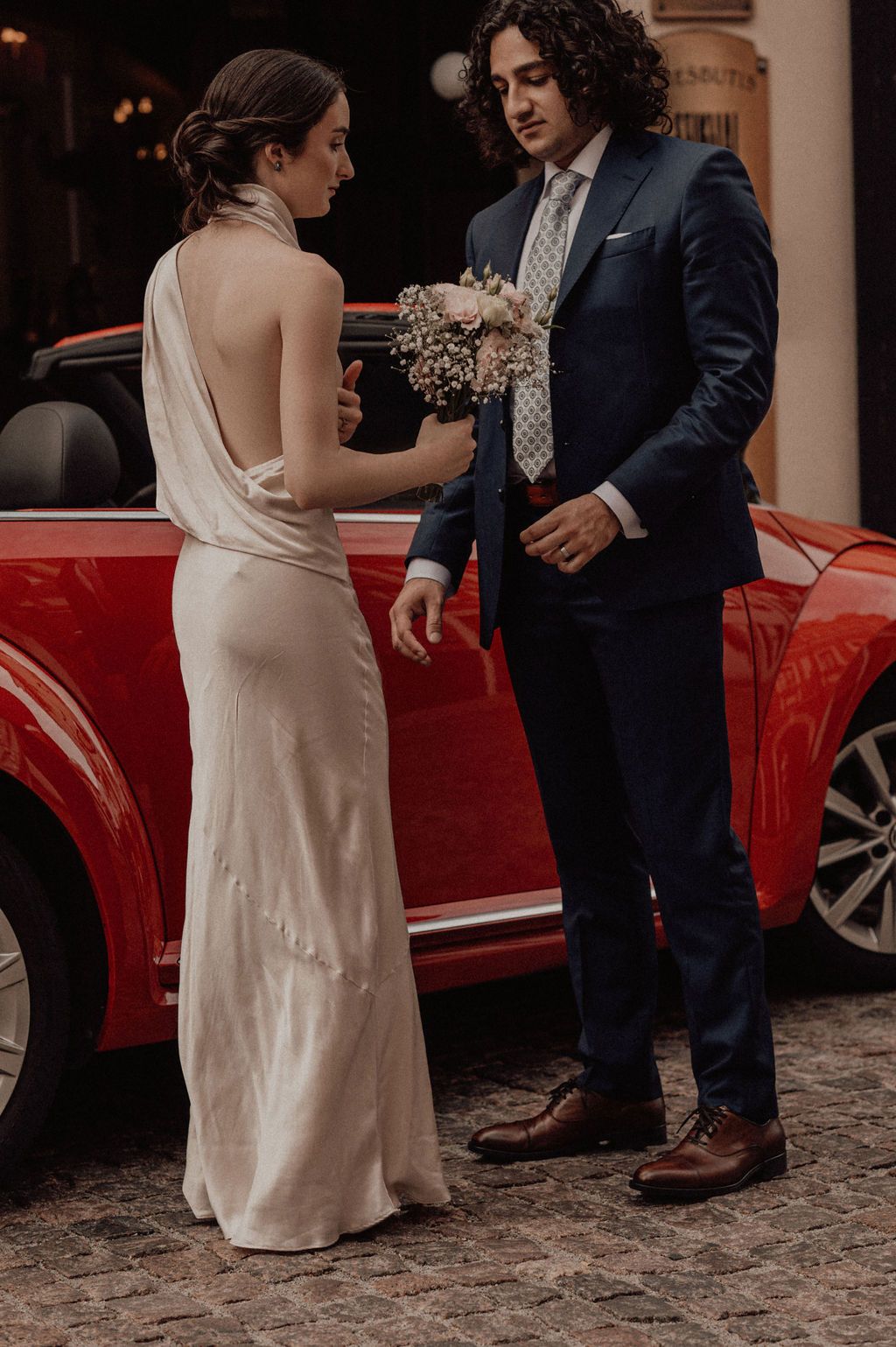 Elegant bride in backless white gown holds bouquet while groom in navy suit stands beside red car on cobblestone street.