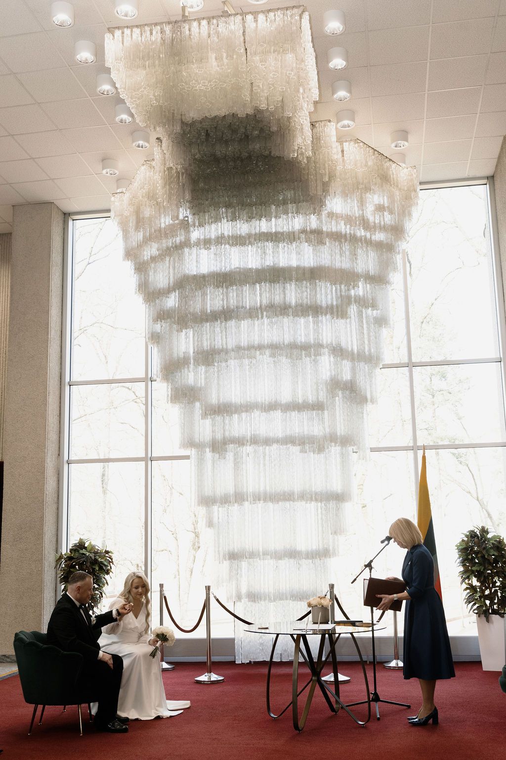 Bride and groom sit during civil wedding ceremony beneath large chandelier with officiant at podium.