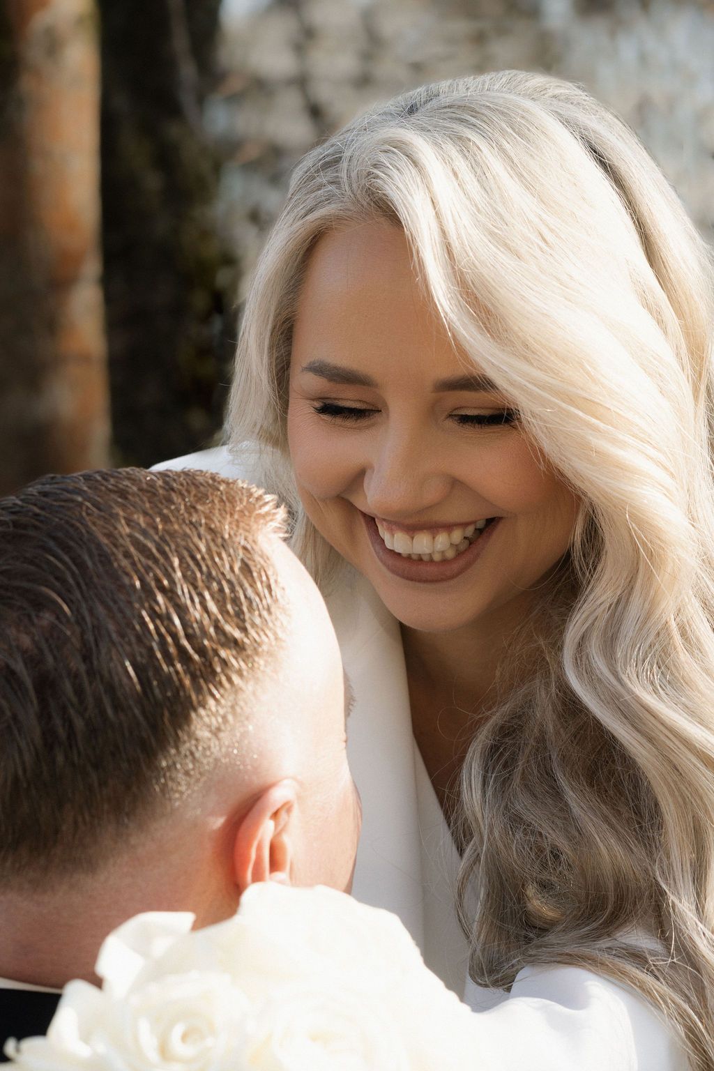 Smiling blonde bride in white dress holds white roses bouquet, sharing a joyful moment with groom outdoors.