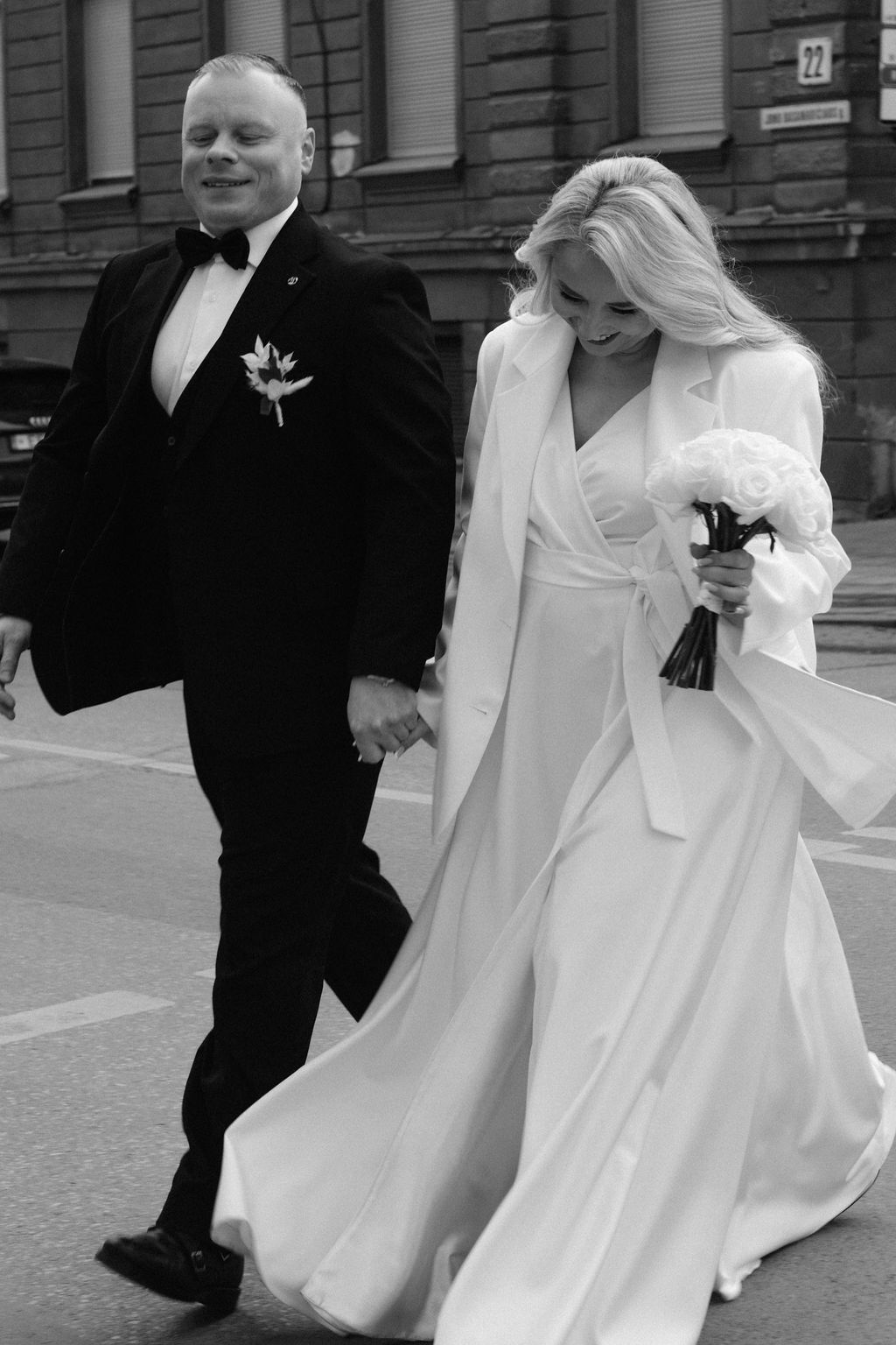 Black and white photo of a smiling bride in white gown holding bouquet walking hand in hand with groom in tuxedo.