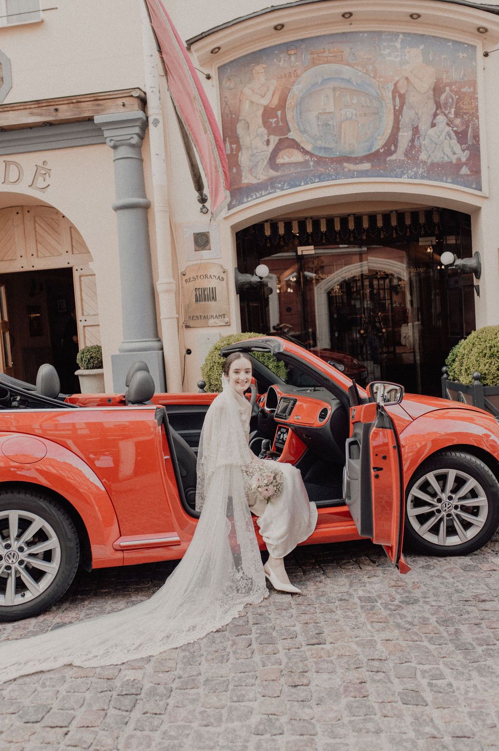 Bride in white wedding dress and veil sitting on red convertible car outside elegant European building.