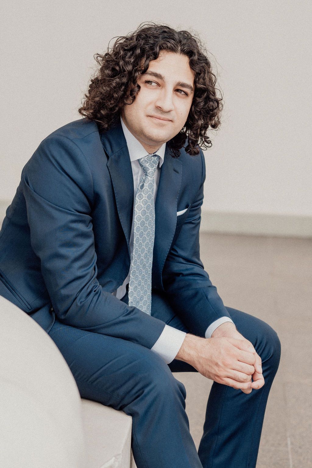A young man with curly dark hair wearing a navy blue suit and patterned tie sits confidently in a modern chair.