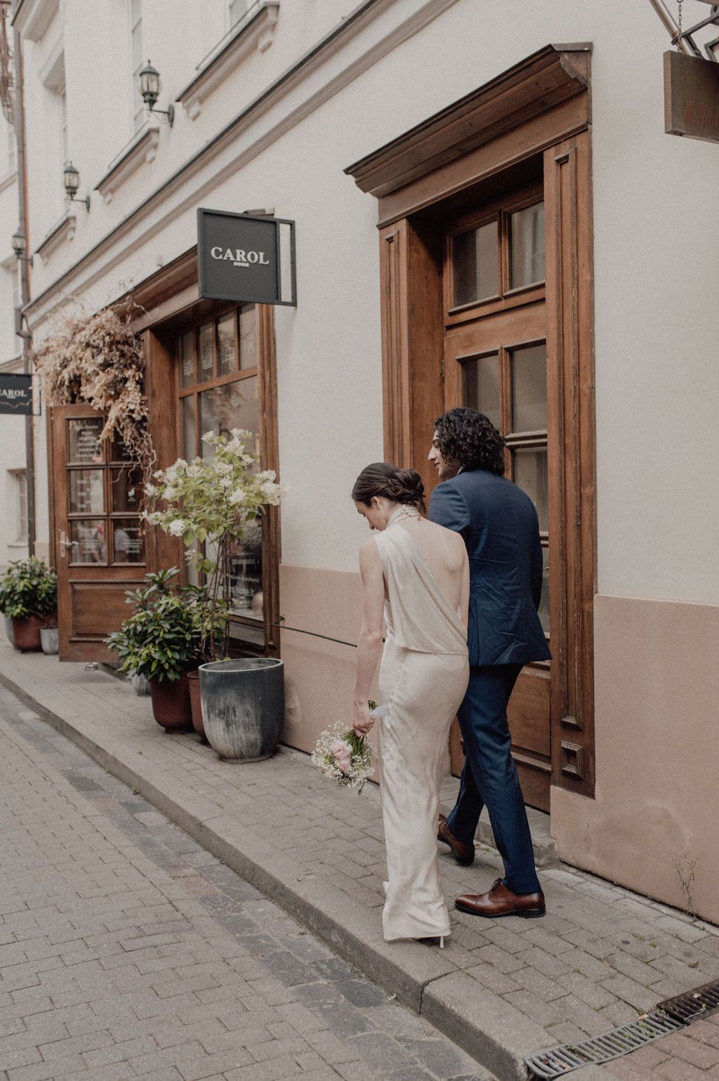 A bride in a white gown and groom in navy suit walk toward a wooden door outside a European cafe.