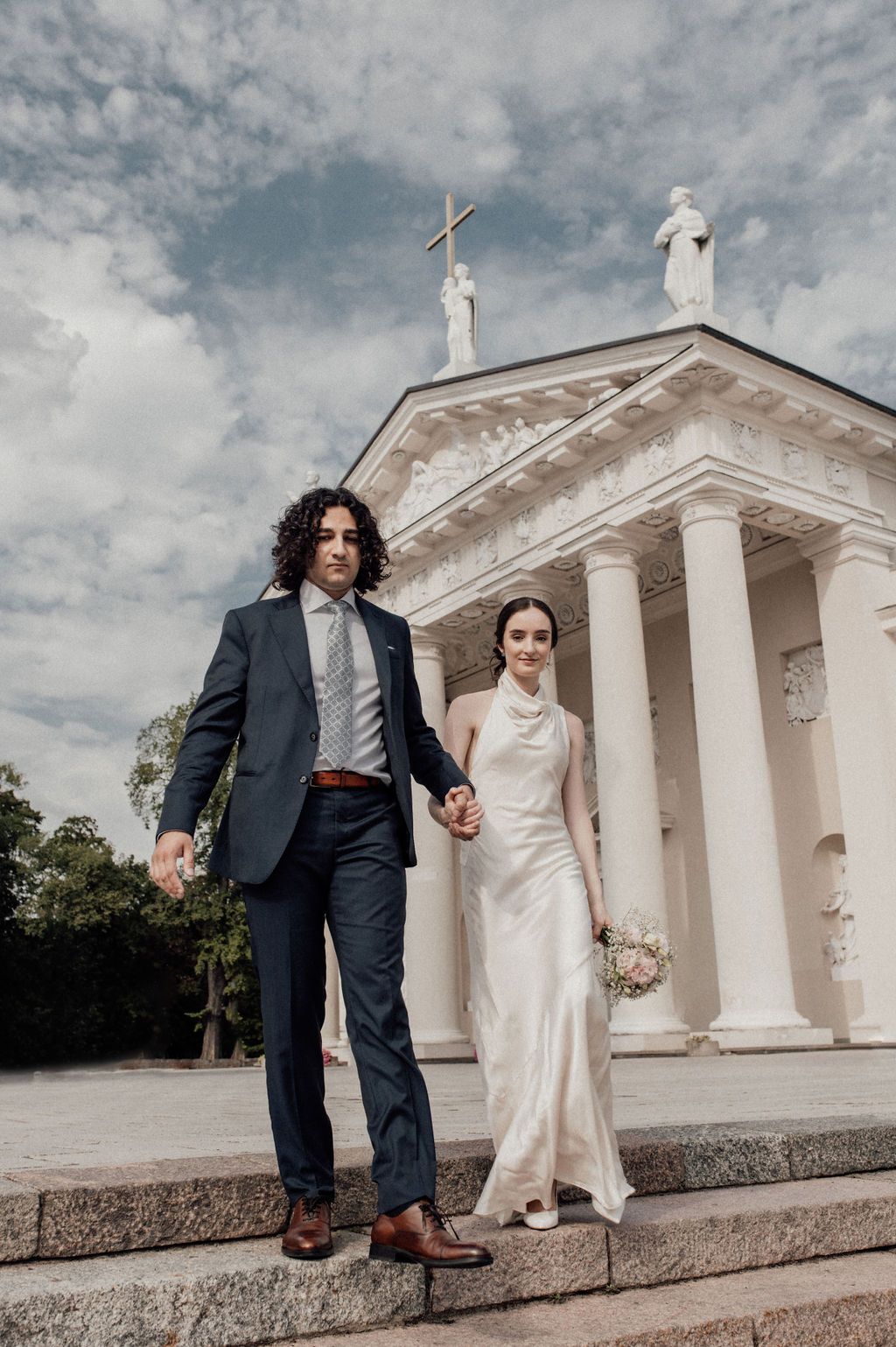 Bride and groom hold hands on church steps, she in white gown, he in navy suit, with a white columned chapel behind.