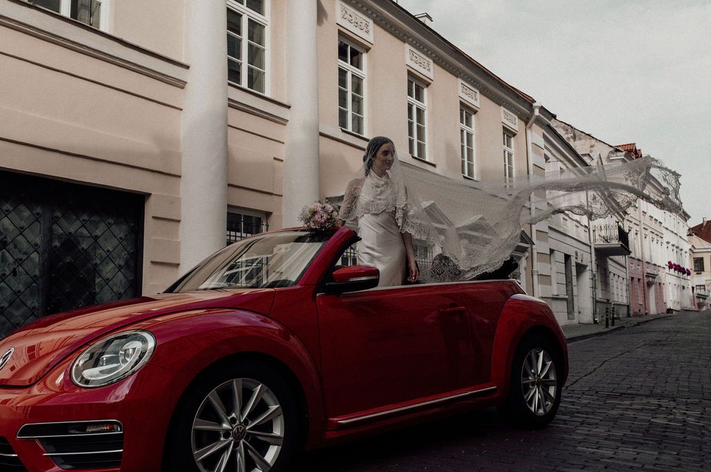 Bride in white gown with flowing veil stands beside a red VW Beetle convertible on a European cobblestone street.