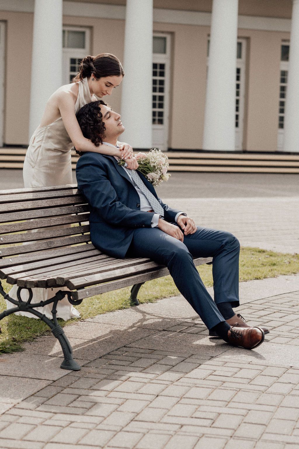 A bride holding flowers stands behind a groom in a navy suit sitting on a park bench outside a white building.