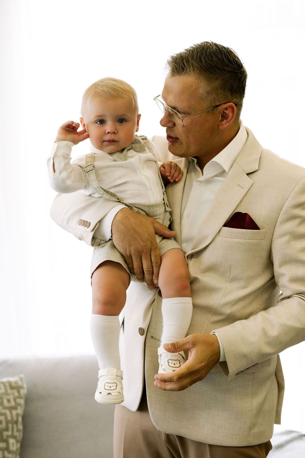 Man in beige suit holding a baby dressed in white outfit, both looking elegant at a formal event.