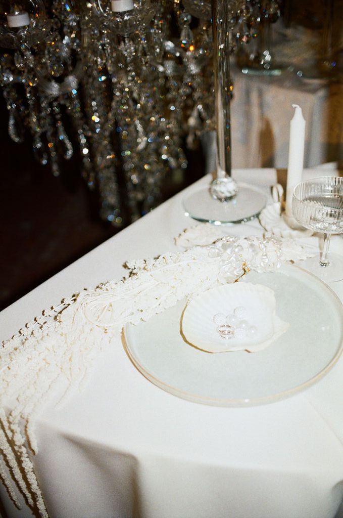Elegant white table setting with crystal chandelier, gold-rimmed plate, white orchid, taper candle, and pearl garland runner.