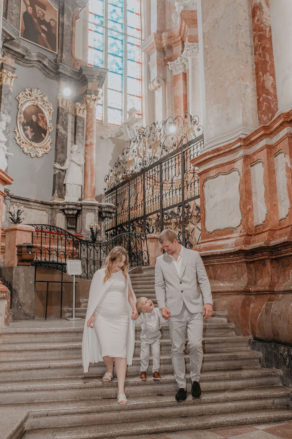 Family in white outfits walking down ornate church stairs, parents holding toddler's hands in elegant baroque interior.
