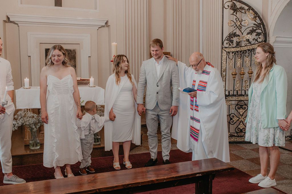 Couple exchanges wedding vows before officiant in elegant church ceremony with family and guests gathered around.