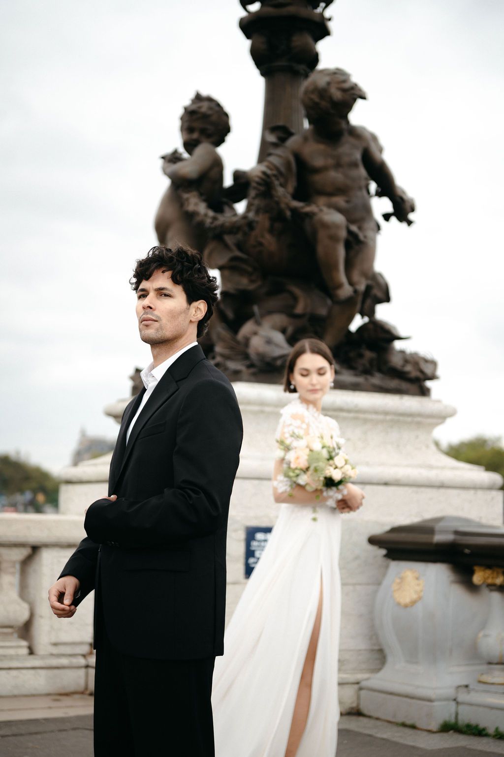 A groom in a black suit stands in the foreground as his bride holds a bouquet near an ornate bronze statue in Paris.