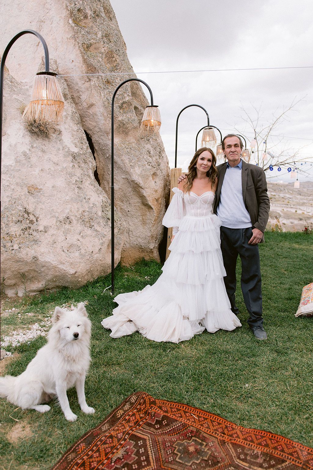 Bride and groom posing outdoors with white Samoyed dog, decorative lamps, and colorful rug in Cappadocia wedding setting.
