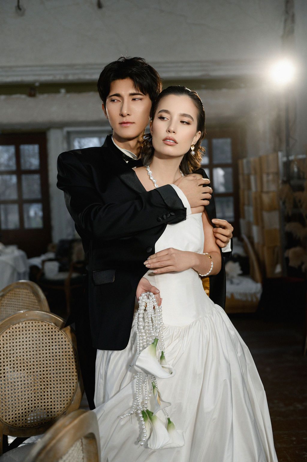 A couple in wedding attire embrace elegantly, groom in black suit, bride in white gown holding a bouquet.