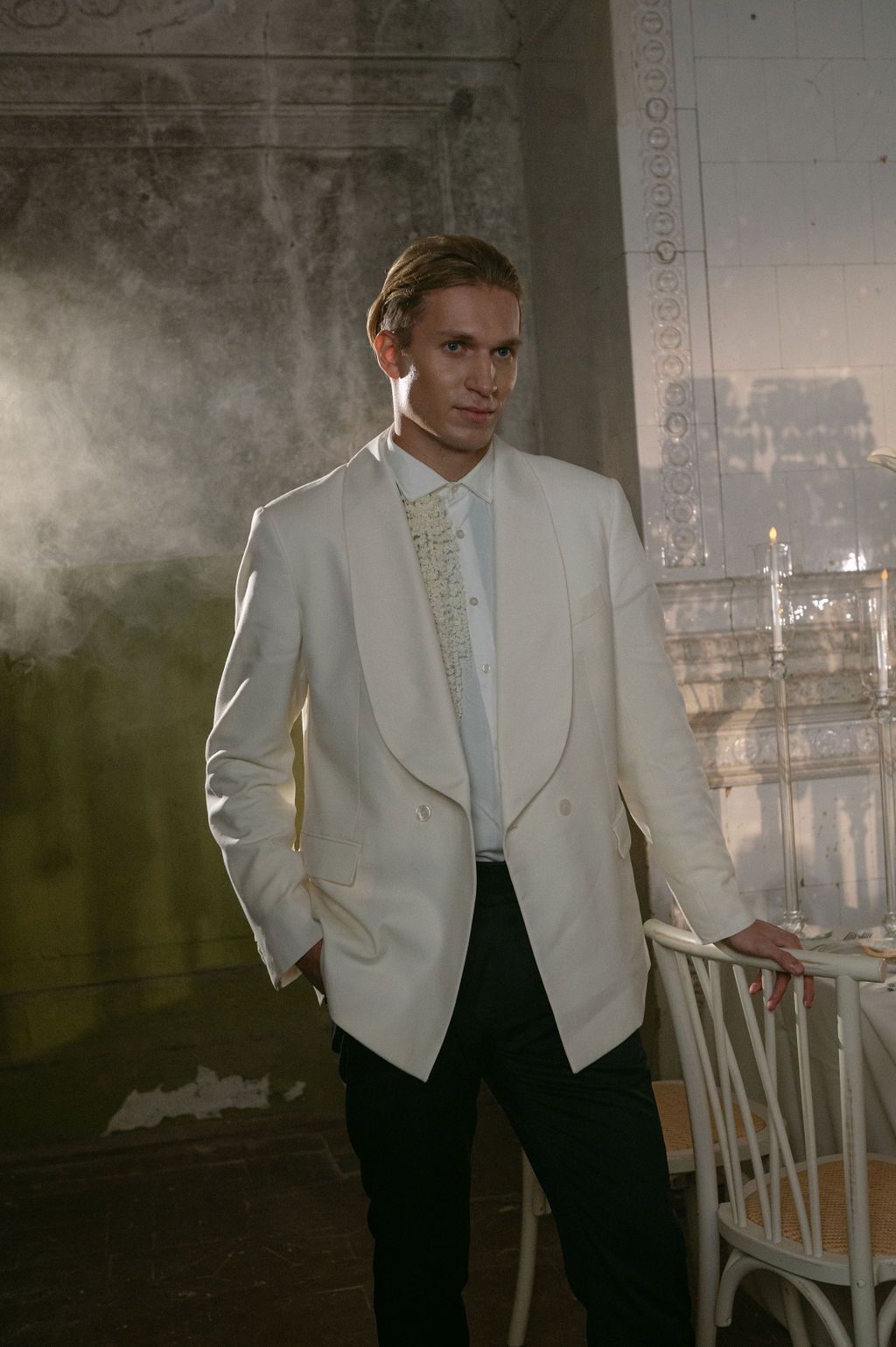Man in white tuxedo jacket with gold tie posing elegantly in a misty, ornate room.