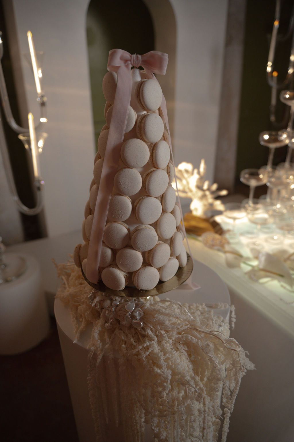 Elegant white macaron tower topped with a pink bow on a wedding cake display with soft bokeh lighting.