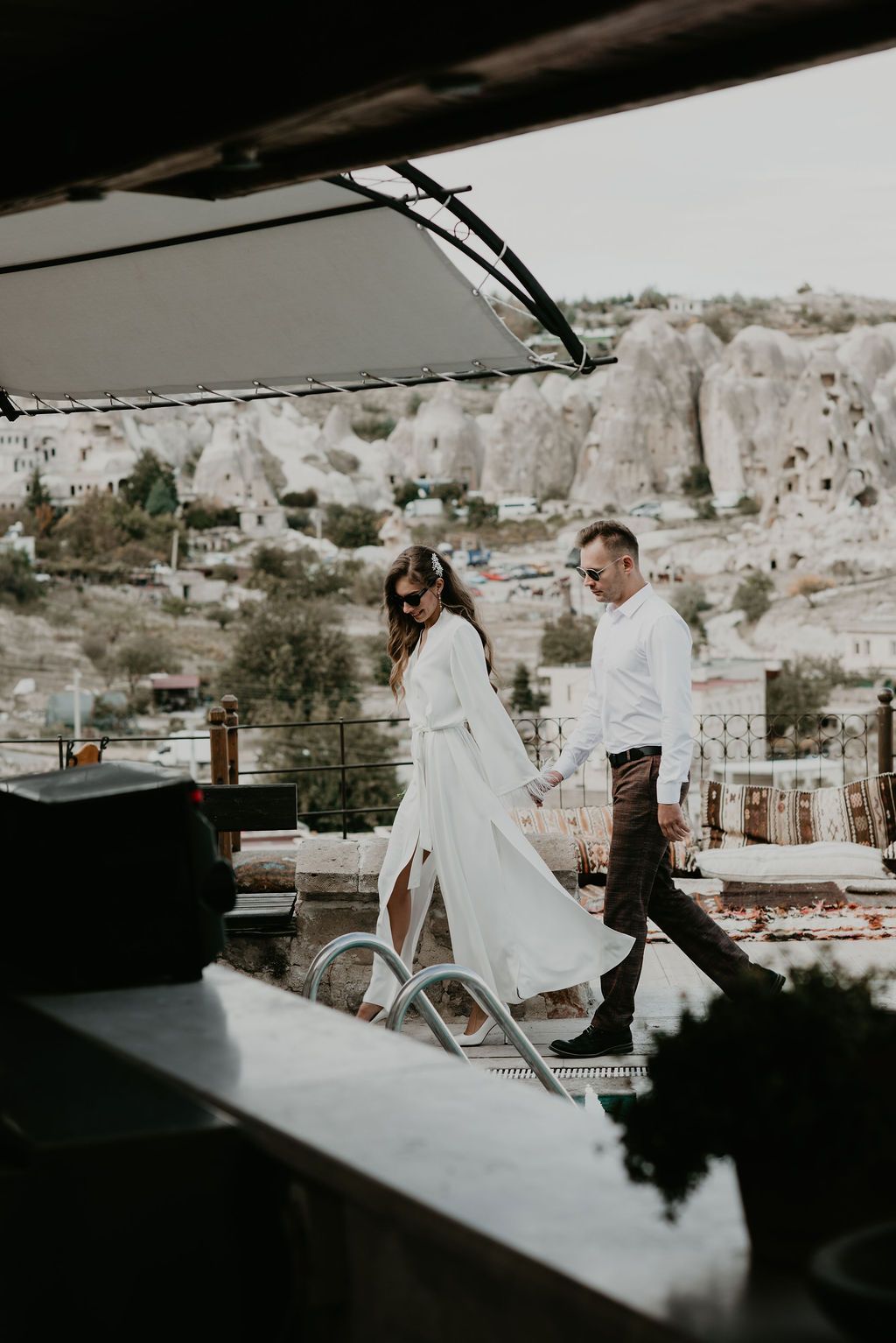 A couple in wedding attire holds hands on a rooftop terrace with Cappadocia cave dwellings in the background.