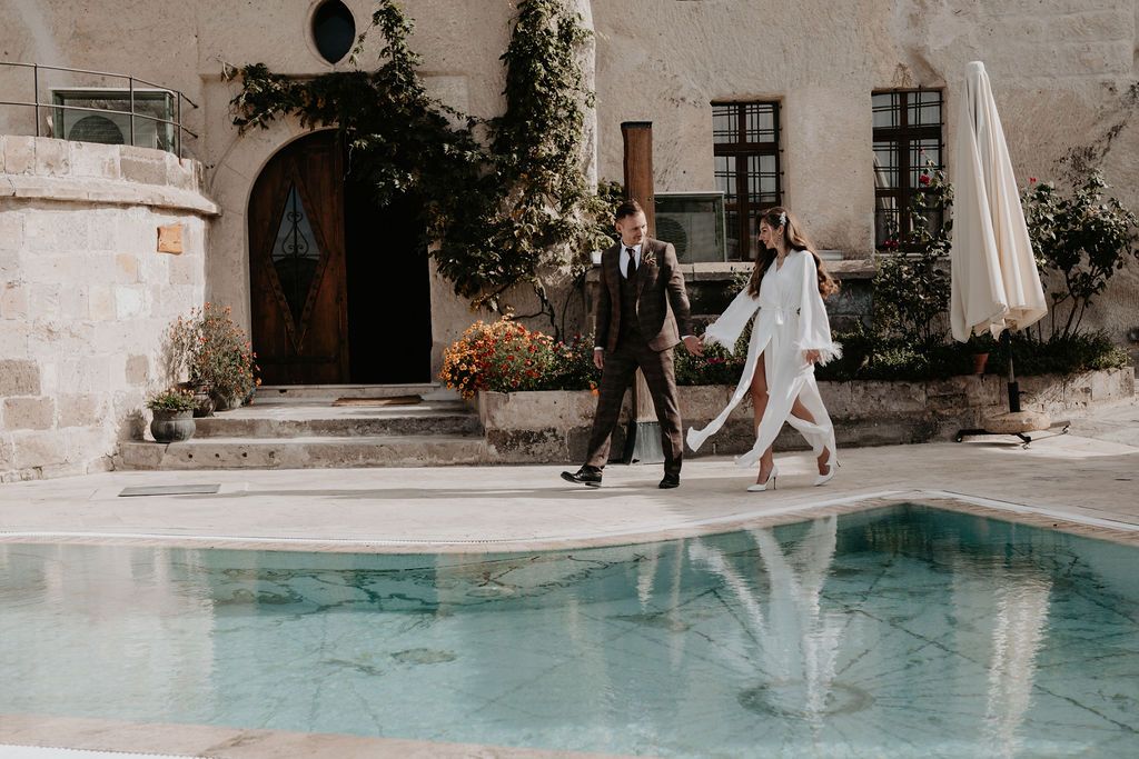 Couple walking hand in hand beside a pool at a stone villa, dressed in elegant wedding attire.