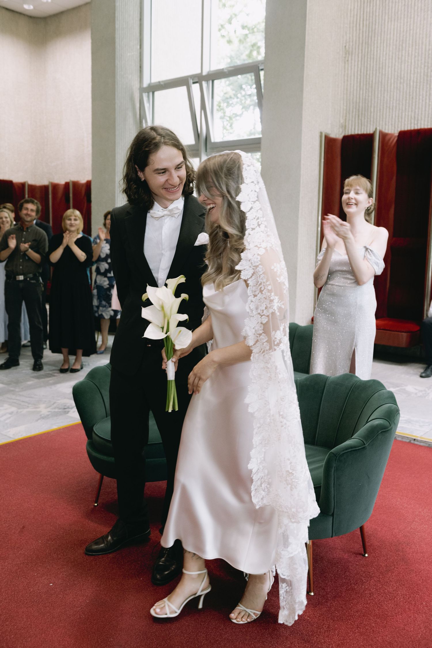 Smiling bride in a satin slip dress and lace veil holds white calla lilies beside a groom in a tuxedo as guests applaud at a civil ceremony.