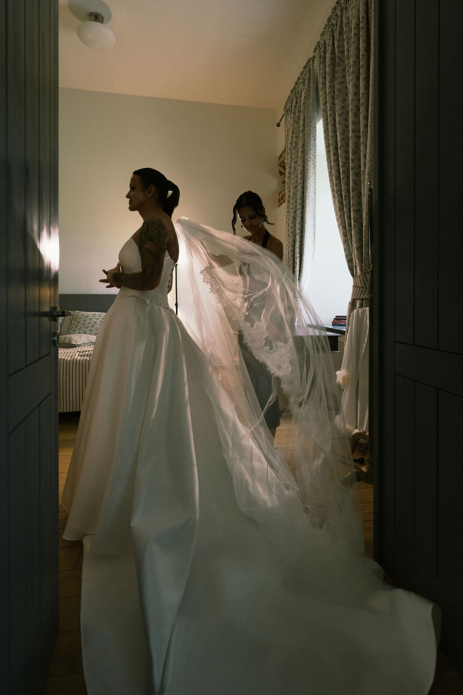 A bride in a white satin gown and long lace-trimmed veil stands in profile in a softly lit room as a helper arranges her veil.