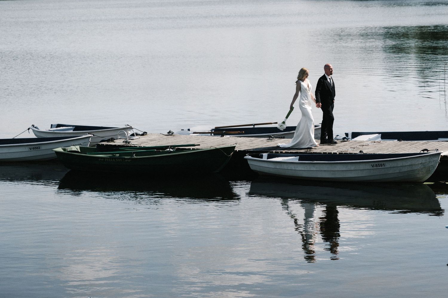 Bride in a white gown and groom in a dark suit stand on a wooden dock among small rowboats, reflected in calm lake water.