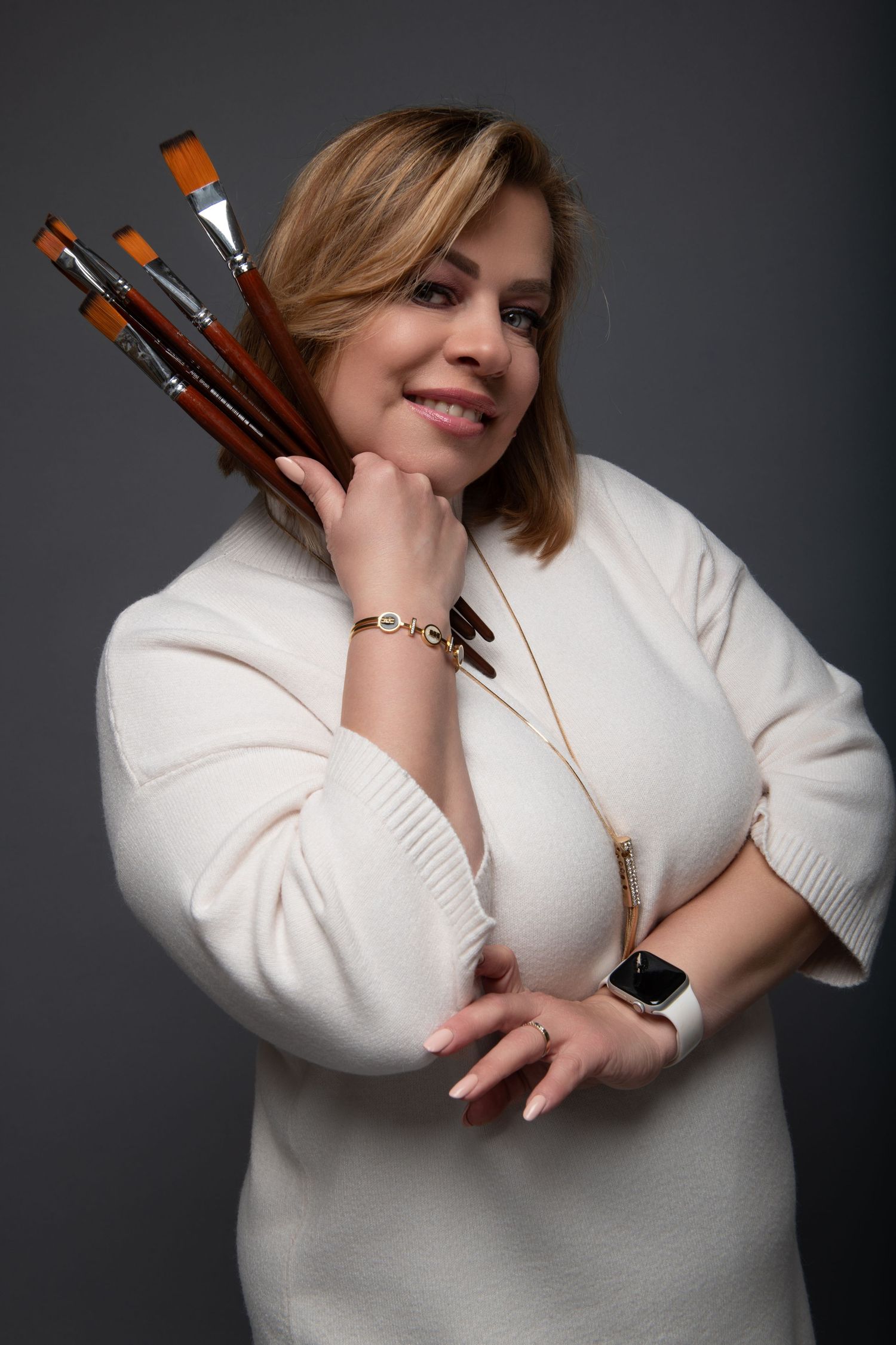 Smiling blonde woman in white sweater holding paint brushes, posing as an artist against gray background.