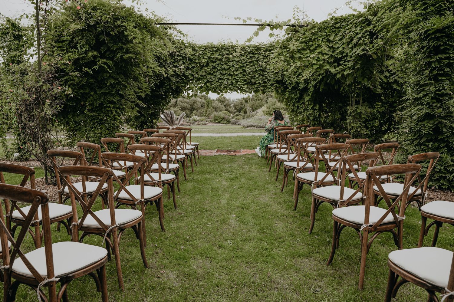 Rows of wooden cross-back chairs with white cushions arranged on a lush green lawn for an outdoor wedding ceremony.