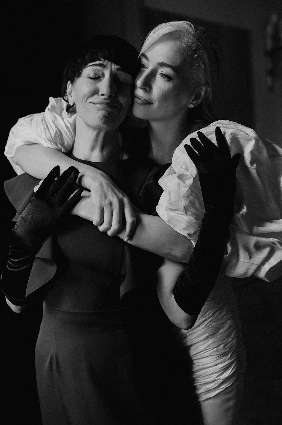 Two women in black and white photo embrace warmly, wearing elegant gloves and contrasting outfits in dramatic lighting.