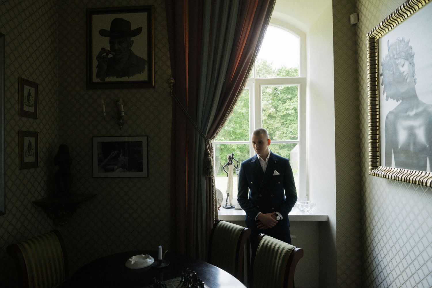 A well-dressed man in a navy suit stands contemplatively by a window in an elegant room adorned with framed portraits.