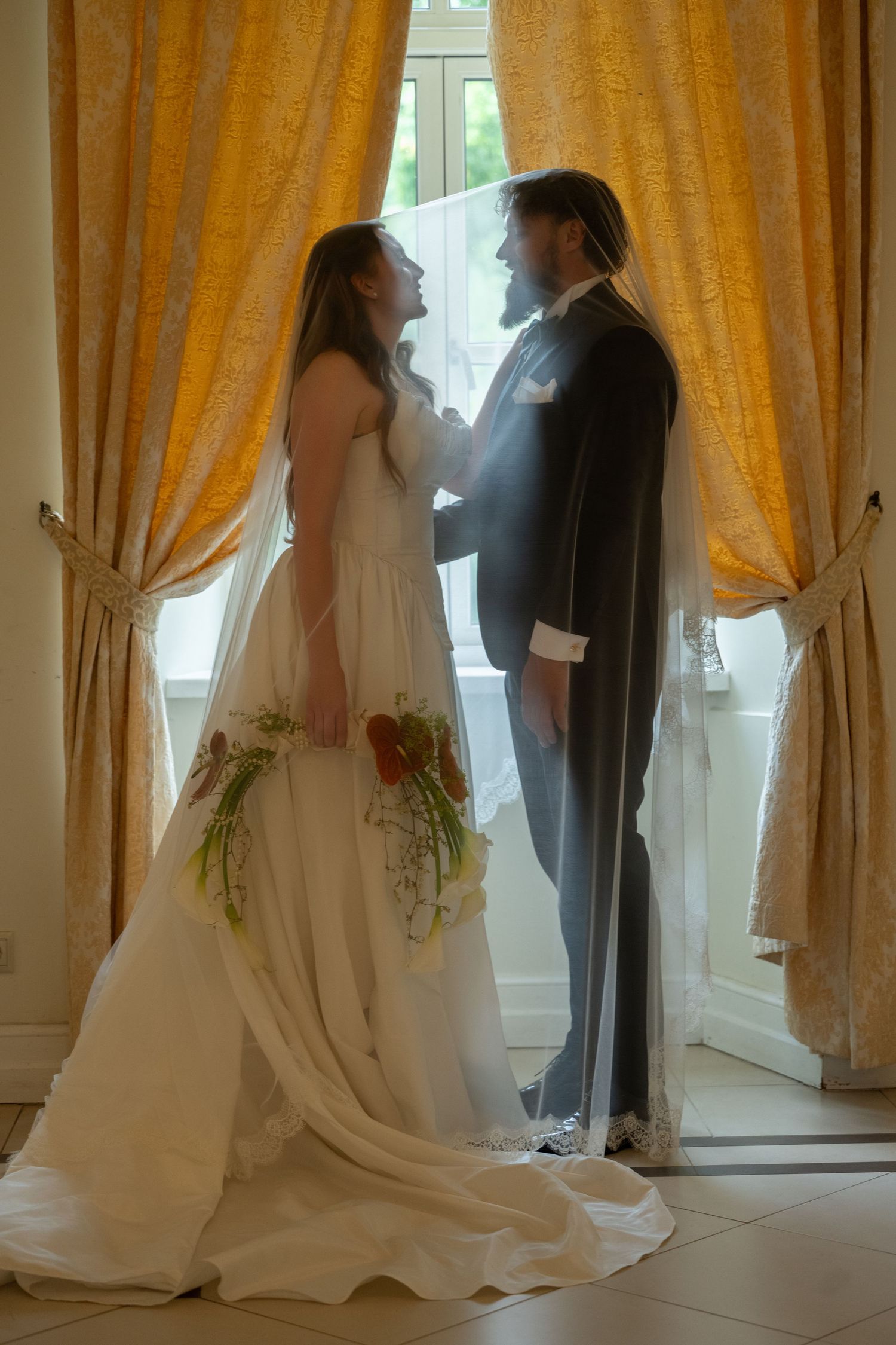 Bride and groom share romantic moment by golden curtained window, bride holding bouquet.