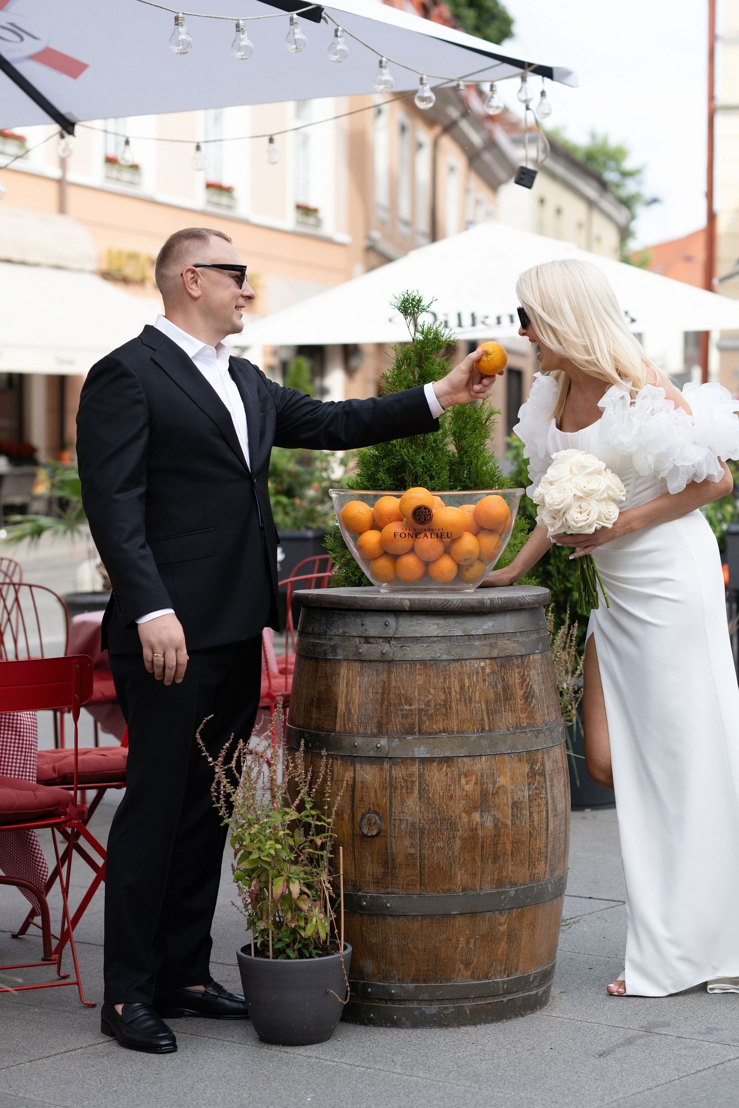 Groom in a black suit and sunglasses picks an orange from a bowl on a wooden barrel beside a bride in a white gown holding a bouquet.