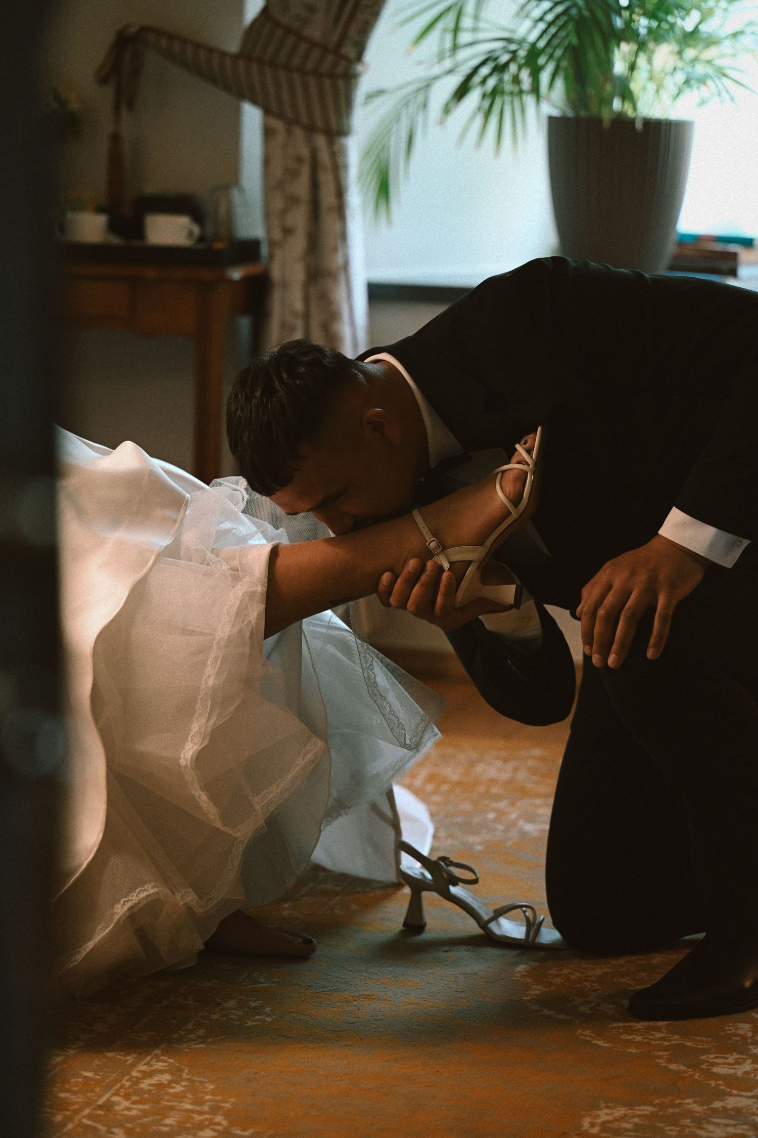 Groom kneels to help the bride put on her strappy high-heeled sandal in a cozy, warmly lit room.