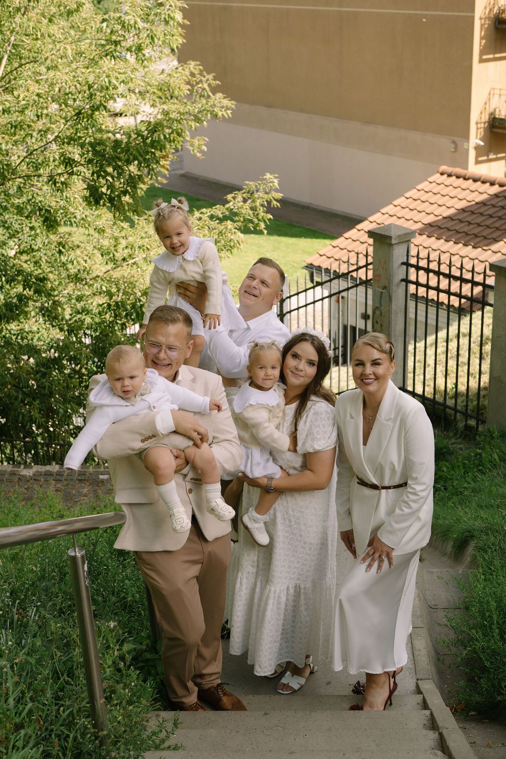 A joyful family group in white and beige outfits poses outdoors on steps surrounded by lush greenery.