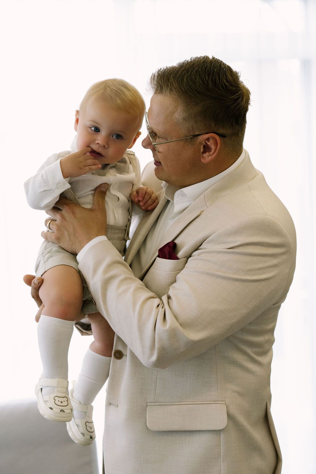 Man in beige suit tenderly holding a baby dressed in white at a formal christening event.