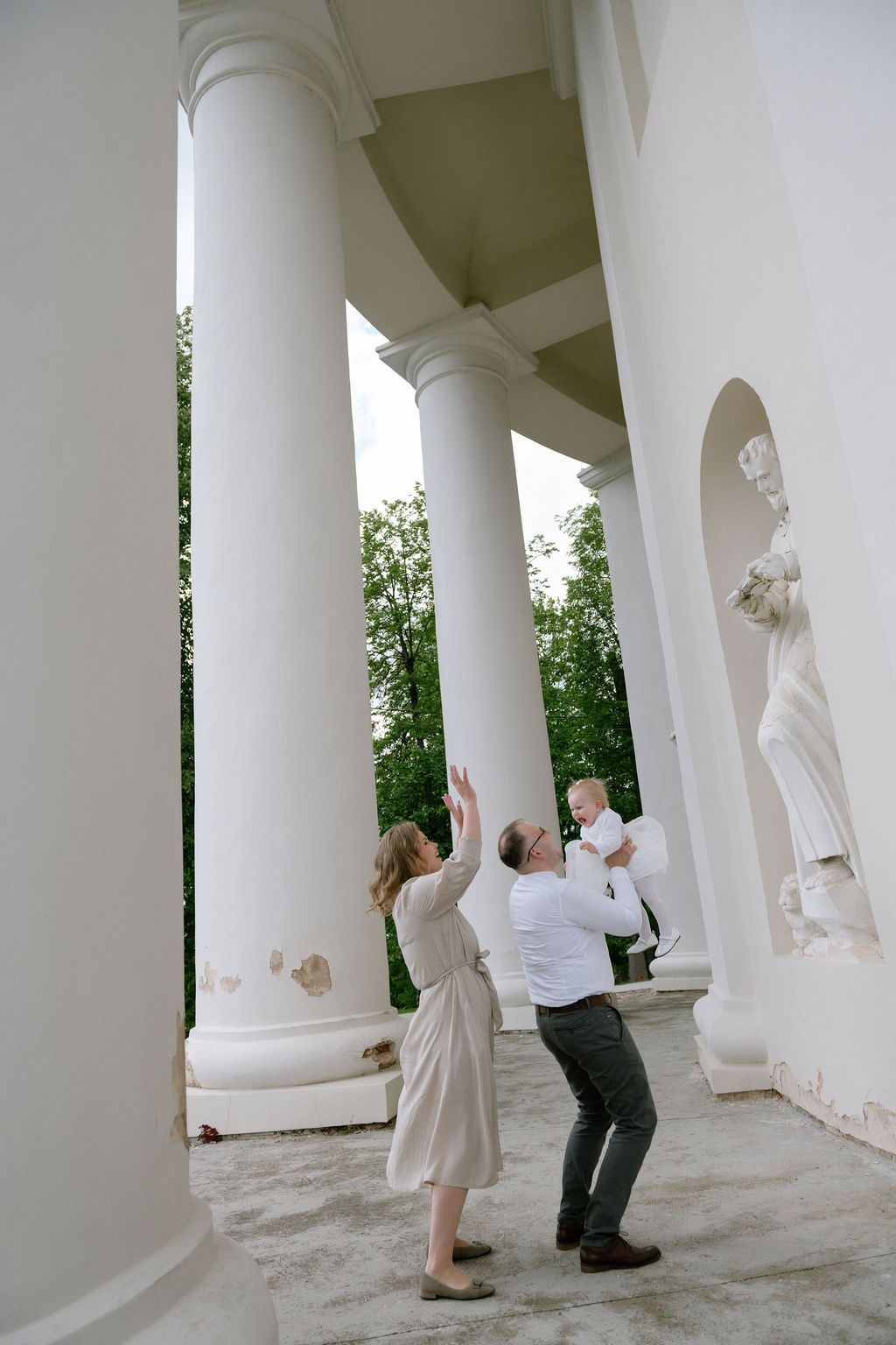 Family with baby playing among white columns of classical architecture building with statues outdoors.