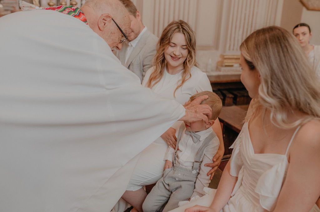 Priest blessing a young boy during a wedding ceremony while two smiling women in white dresses look on warmly.