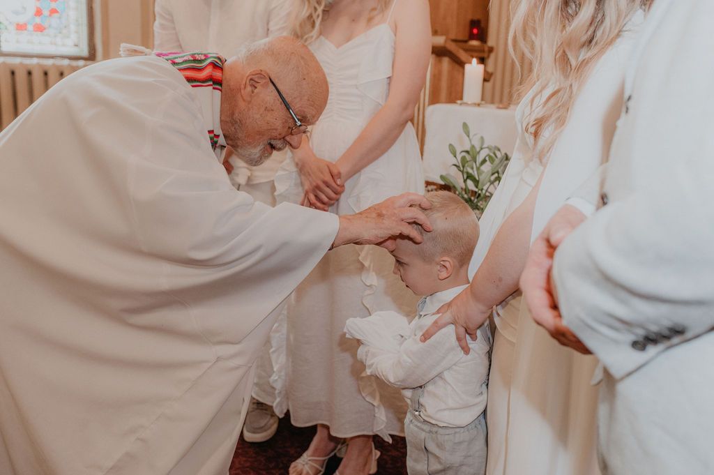 Priest performing a baptism ceremony on a baby dressed in white, with family members gathered around in a church.
