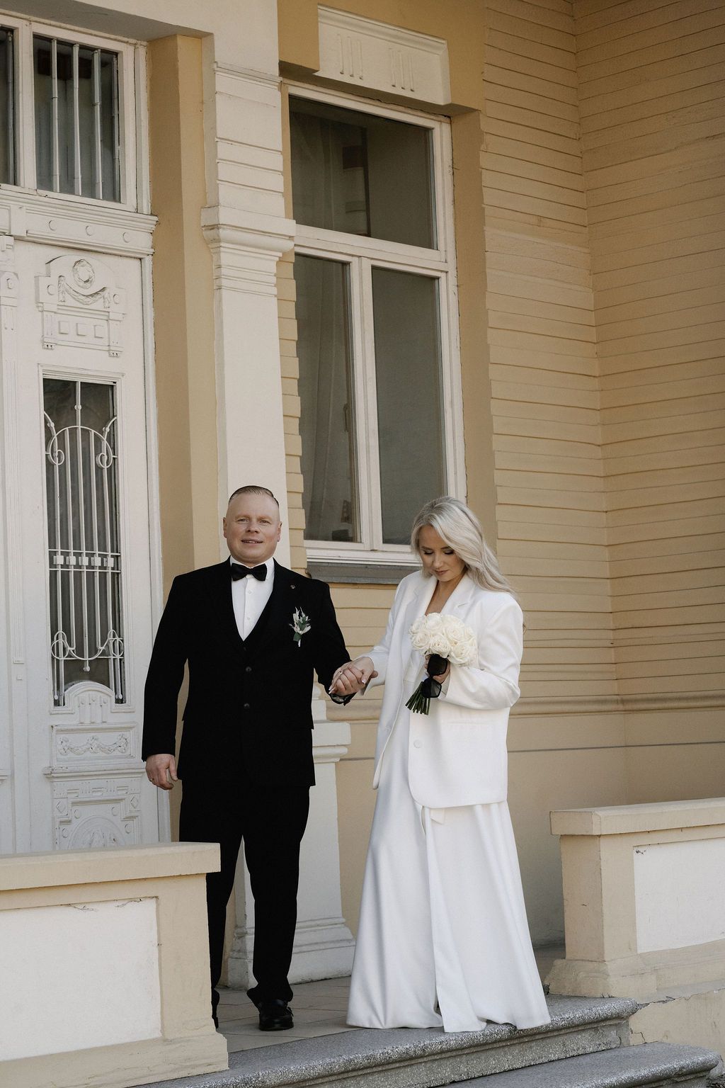 Elegant bride in white suit holding bouquet walks with groom in black tuxedo outside a classic building.