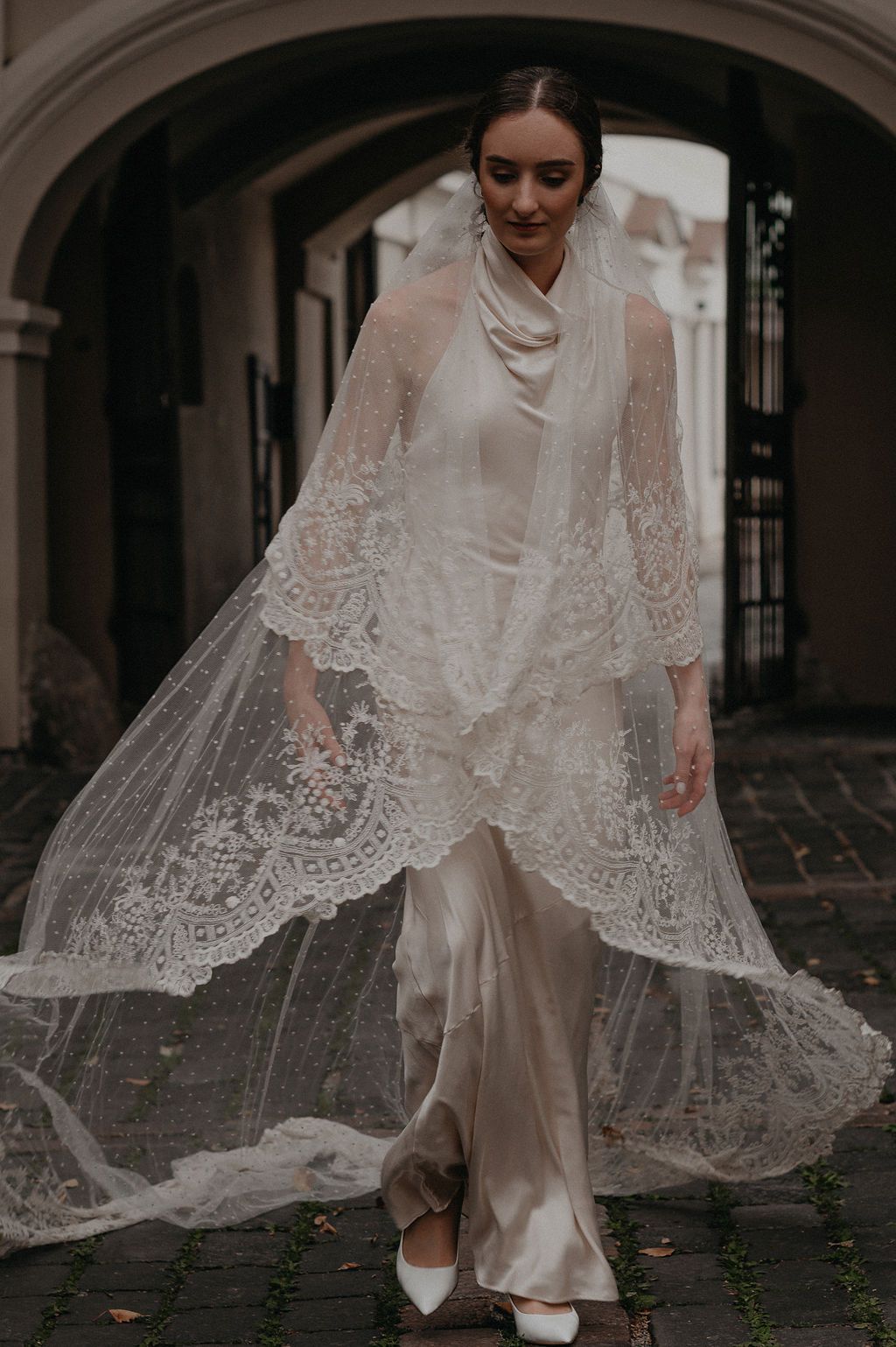 Bride in satin gown with flowing lace veil walks through stone archway, looking down elegantly.