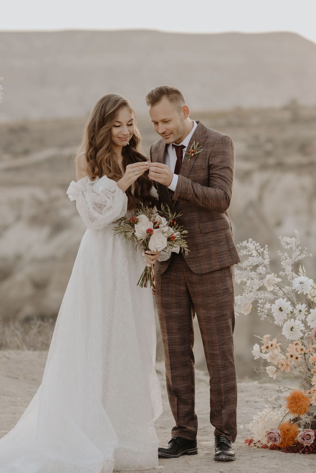 Bride in white gown and groom in brown plaid suit exchange rings outdoors with floral arrangements nearby.